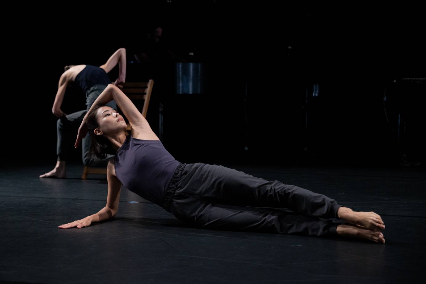 two female dancers, one in the foreground in a grey leotard and semi-fitted sweats holds an angular pose lying on her side the floor,the dancer in the background, dressed similarly sits on a chair huddled over her back faces us